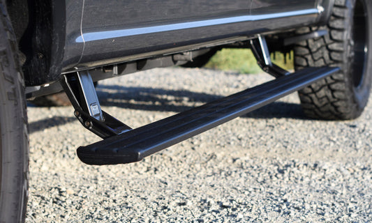 Close-up of a vehicle’s AMP Research PowerStep Smart Series retractable side step bar for the 15-20 Chevy/GMC Silverado 2500-3500 and Sierra 2500-3500 Crew & Double Cab, showcasing its black textured surface and electric-powered running board. The mechanism stands out against a gravel background, with hints of grass visible beyond.