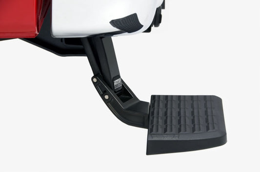 The AMP Research 1999-2016 Ford F-250/350/450 BedStep in black extends from the rear bumper of a red and white pickup truck, featuring a non-slip textured surface for grip and a sturdy, foldable design.