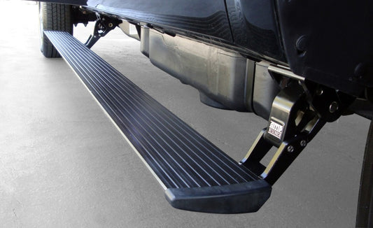 The close-up highlights an AMP Research 2015-2016 Chevy Silverado 2500/3500 Double/Crew Cab PowerStep Plug N Play - Black, resembling an automatic running board, extended from the side of the vehicle. Its ribbed surface ensures traction, while the metal mechanism underneath offers robust support against the smooth concrete backdrop.