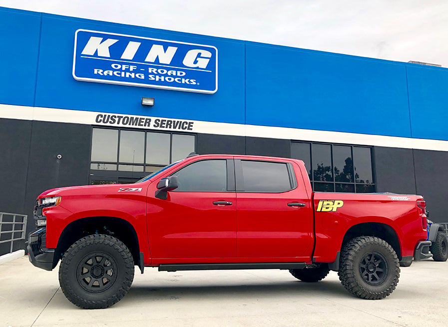 A red pickup truck with large off-road tires is parked in front of a building with a blue and white sign that reads "KING Off-Road Racing Shocks." The building has a "Customer Service" sign above the entrance.