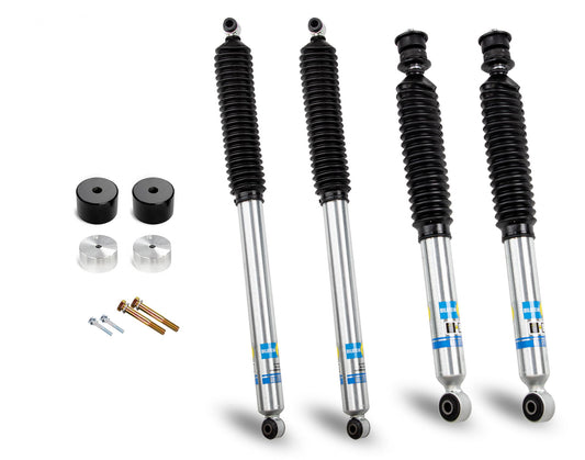 Image of four Cognito 2-Inch Economy Bilstein shocks lined up vertically alongside a set of black rubber bushings, aluminum spacers, and bolts with washers on the left side. The shocks feature silver bodies with black dust boots and blue and yellow logos near the bottom, making them ideal for upgrading a 2005-2016 Ford F250/F350 4WD truck leveling kit.