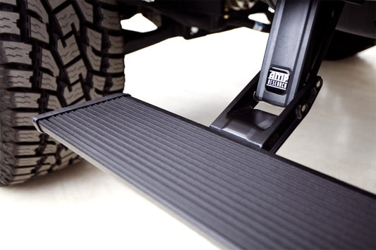 Close-up of an AMP Research 21-23 Ford Bronco PowerStep Xtreme - Black retractable truck step with a textured surface, extending from under the vehicle next to a large tire. Designed for extreme environments like sub-zero winters, the step prominently features the "AMP Research" label. The background is blurred, drawing attention to the step and tire.