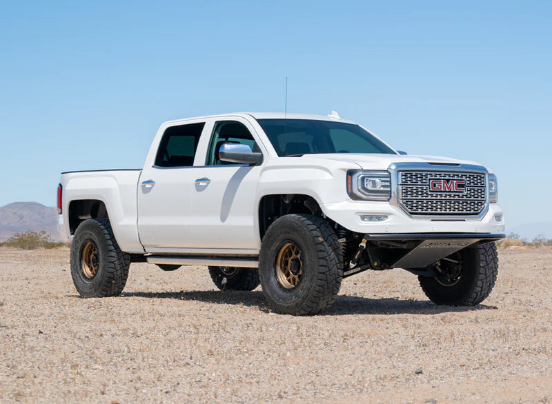 A lifted white GMC pickup truck is parked in a desert landscape with a clear blue sky. The truck features an aftermarket suspension lift, large off-road tires, and a custom front bumper. The terrain is sandy and barren with distant mountains visible.