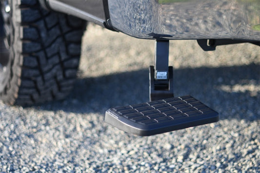 Close-up of the AMP Research 21-22 Ford F150 BedStep, a retractable step designed to assist with vehicle entry. The black step features a textured surface for enhanced grip, making it ideal for cargo loading, set against a backdrop of gravel. Note that it is not compatible with dual exhaust systems.