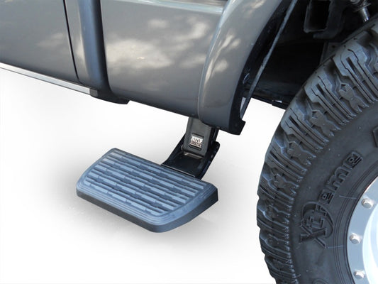 Close-up of an AMP Research 2009-2014 Ford F-150 All Beds BedStep2 in black next to a large tire. The retractable truck step is extended, featuring a textured surface for grip, and is seamlessly attached to the side of a gray vehicle.