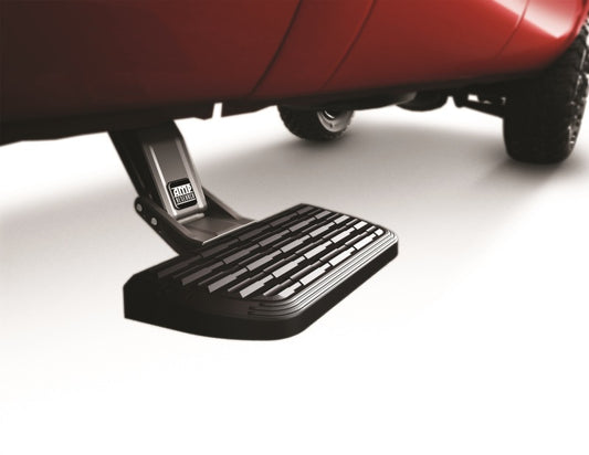 A close-up shows the AMP Research 1999-2016 Ford F-250/350 All Beds BedStep2 in black mounted below a red vehicle. This retractable truck step, designed by AMP Research, features a textured surface for enhanced grip and is extended outward. The step's dark color contrasts with the surrounding white floor, emphasizing its practical design.