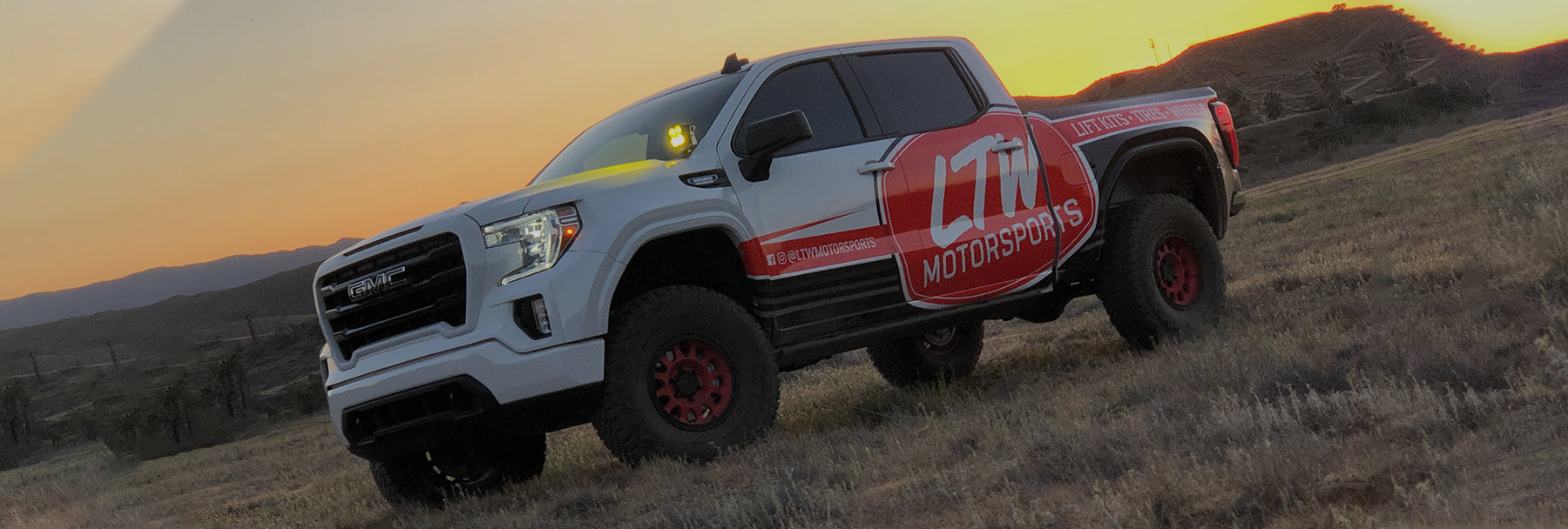 A white pickup truck with red and black detailing and a large "LTN Motorsports" logo on the door is parked on a grassy field at sunset. The truck has a robust build with off-road tires and a light bar mounted on the roof. Mountains and a tree are in the background.