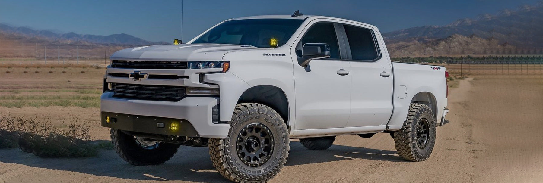 A white Chevrolet Silverado truck is parked on a dirt road in a desert-like environment with mountain ranges visible in the background. The truck has large off-road tires and a rugged appearance. The sky is clear and blue.