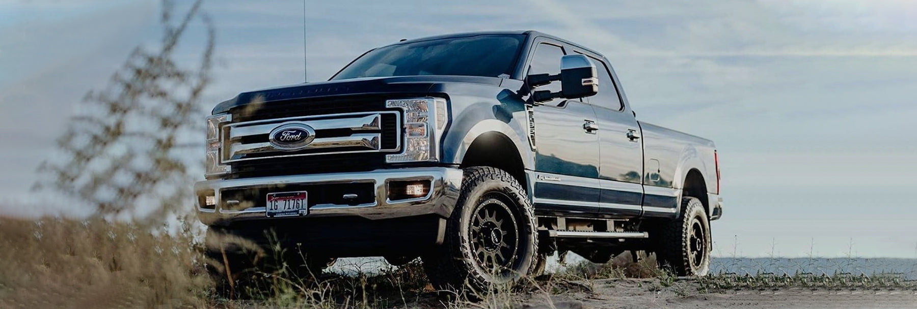 A black Ford pickup truck with large off-road tires is parked outdoors on a dirt road. The sky is partly cloudy, and tall grass is in the foreground. The vehicle's grill and headlights are prominently visible.