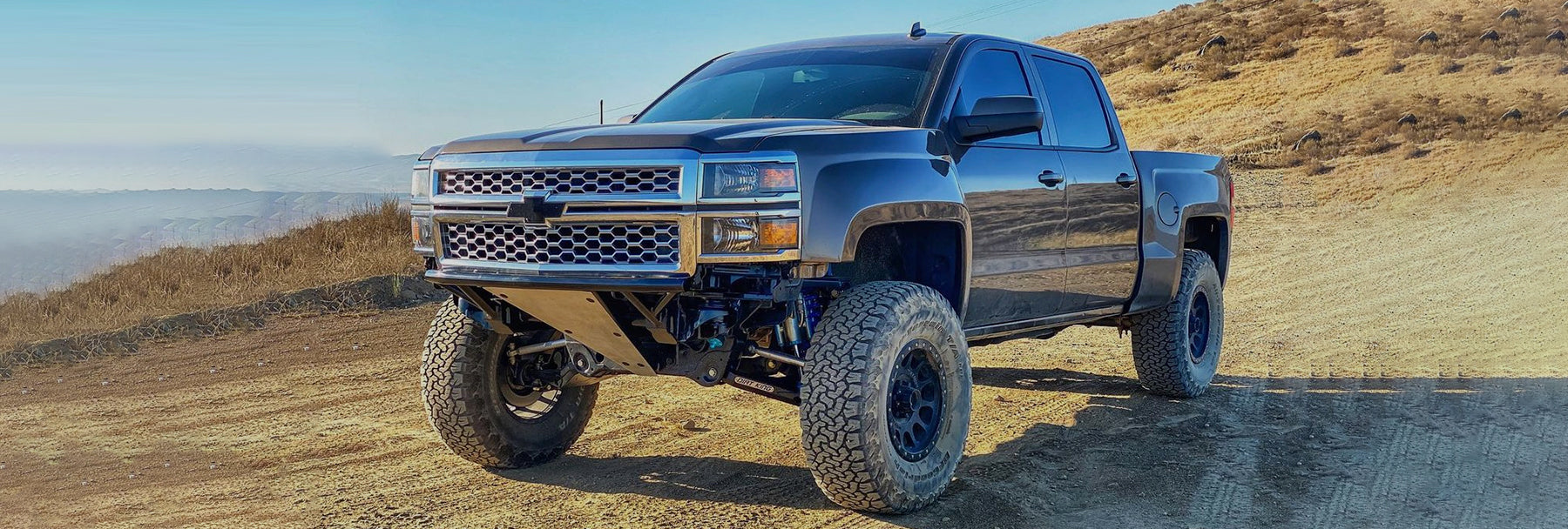 A lifted black Chevrolet Silverado truck is parked on a dirt trail with a hilly, dry landscape in the background. The truck has large tires and a rugged appearance suitable for off-road driving. The sky is clear with a hint of haze.