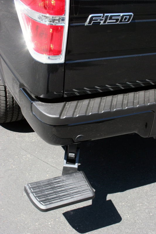 Close-up of the rear of a black truck fitted with the AMP Research 2006-2014 Ford F150 BedStep in black, which features an extended, non-slip bumper step designed for easy access to the truck bed. The vehicle is parked on a paved surface.
