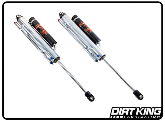 The image showcases two custom-built off-road racing shocks by Dirt King Shocks. These Dirt King Bed Cage Spec Fox 16" Triple Bypasses feature chrome-plated bypasses with attached reservoirs, complemented by black detailing and the Dirt King logo. The image is set against a white background framed by a thick black border, with the Dirt King Fabrication logo positioned in the bottom right corner.
