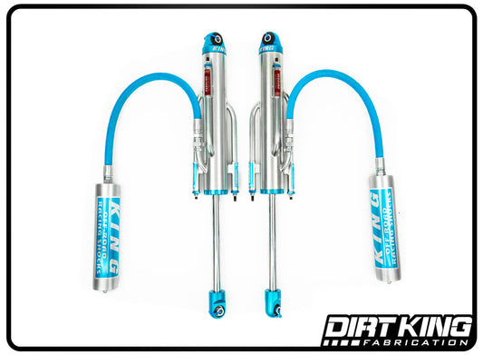 Two Dirt King 19-23 GM 1500 Long Travel Spec King Triple Bypasses are displayed, featuring silver and blue shock absorbers with remote reservoirs connected by blue hoses. The bypass shocks proudly display the DIRT KING SHOCKS logo in blue text on both the shock absorbers and the reservoirs. A Dirt King Fabrication logo appears in the bottom-right corner.