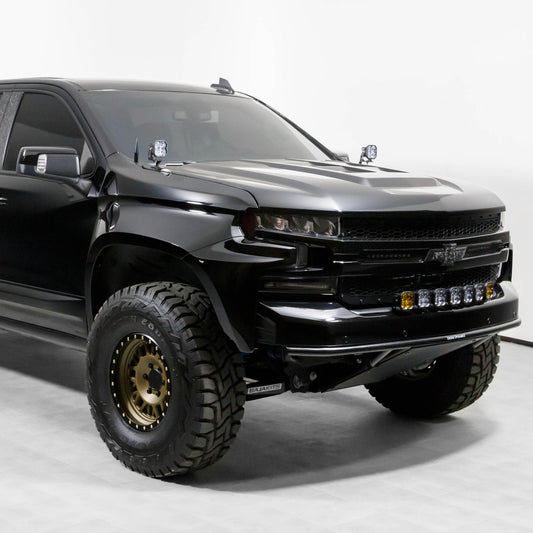 A side-front view showcases a black off-road truck equipped with 2019-2021 CHEVROLET SILVERADO FENDERS by ADV Fiberglass, featuring large rugged tires, additional front bumper lights, and side mirrors with integrated turn signals. The truck boasts a sleek and aggressive design with a matte finish, set in a well-lit studio environment.