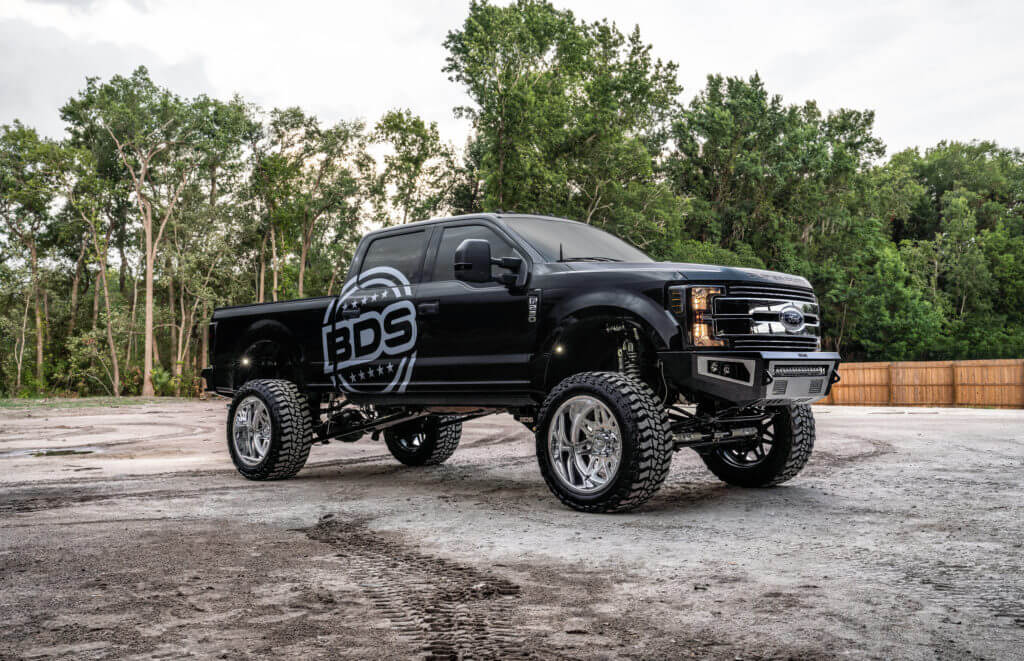 A black pickup truck with large, shiny chrome wheels and a lifted suspension is parked on a gravel surface. The truck has "BDS" logo decals on the doors. There are trees and a wooden fence visible in the background.