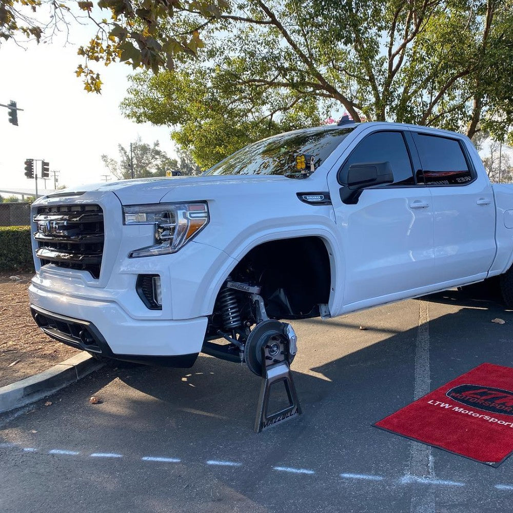 A white pickup truck is missing its right front wheel, with the axle propped up on a jack. The truck is parked in a lot next to a tree, with a red rectangular mat on the ground nearby.