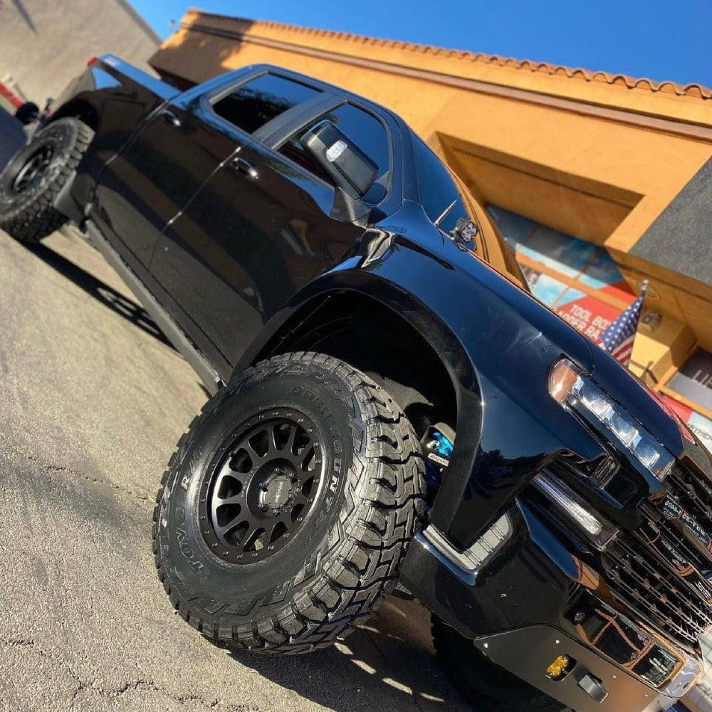 A lifted black pickup truck with large off-road tires is parked on a sunny day in front of a tan building with a tiled roof. The truck's grill and headlights are prominently displayed, and the angle of the shot emphasizes its height and rugged appearance.